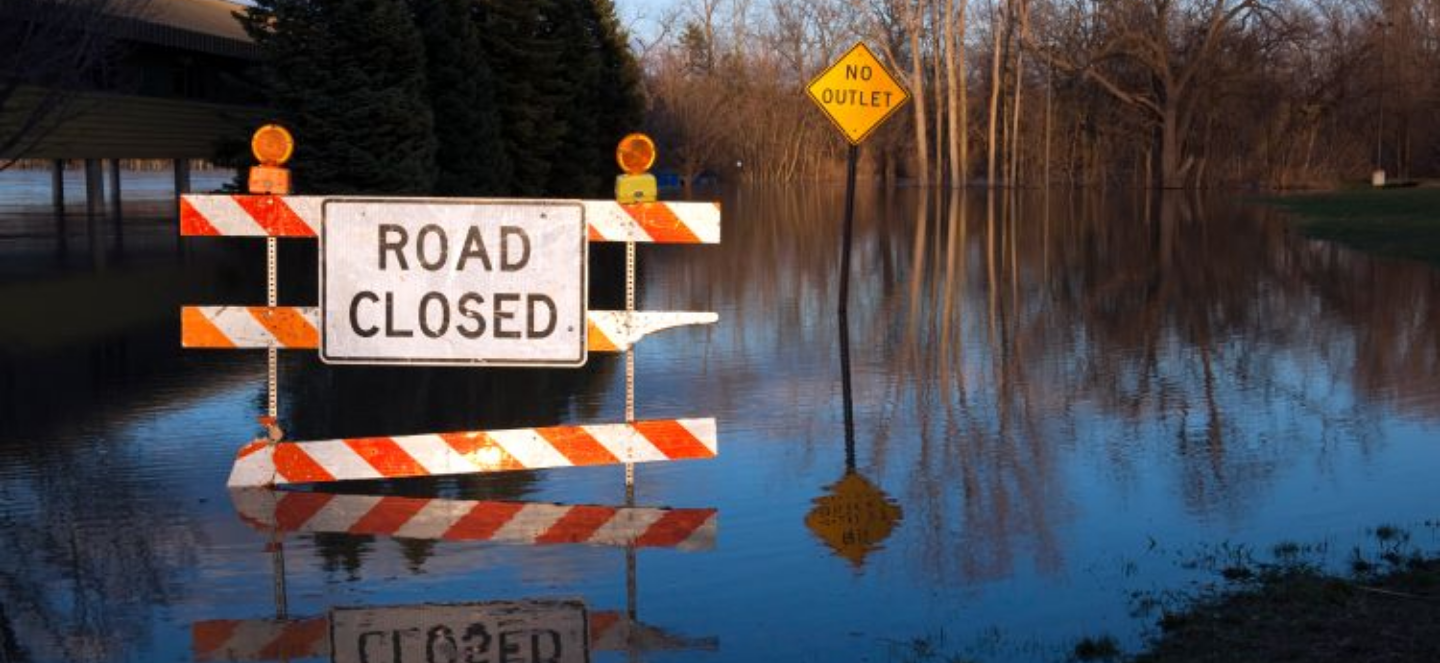 An orange and white road closed barrier sign stands in floodwater on a submerged road, with a no right turn sign partially visible and flooded trees in the background.