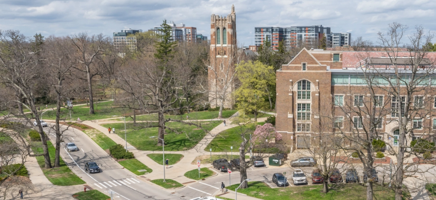 Aerial view of a university campus showing a brick academic building in the foreground, a tall bell tower, tree-lined walkways, and surrounding campus buildings in spring.