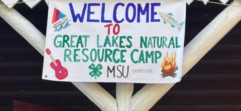 A handmade banner reading “Welcome to Great Lakes Natural Resource Camp” with drawings of a fish, guitar, sailboat, campfire, and the MSU Extension 4-H clover logo.
