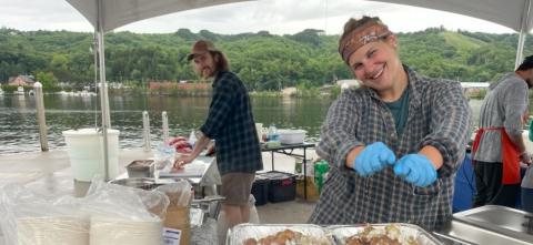 A smiling person wearing gloves prepares food under a canopy by the water, while others work in the background with mountains and trees across the lake.