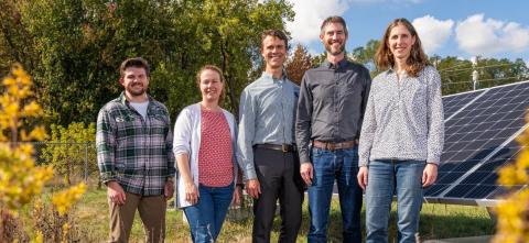 Five people stand outdoors near solar panels with trees and blue sky behind them.