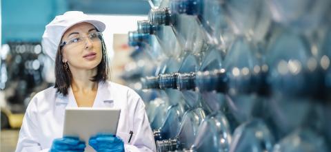 A researcher in protective gear examines industrial equipment while holding a tablet.