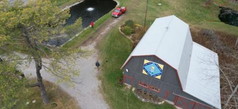 Aerial view of a barn beside a pond and gravel driveway with people and a red truck nearby.